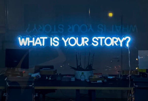 in neon lights the text says What is your story? in the window of a darkened office