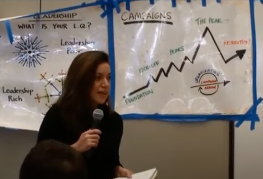 woman holding microphone speaking. There are posters with graphs on the wall behind her.