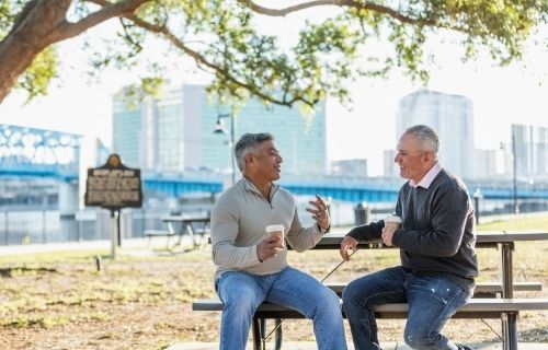 two men sitting on park bench and table having a coffee and a chat with buildings in the background